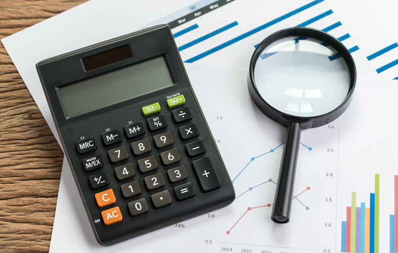 Business perfomance review or KPI key point indicator estimation concept, calculator with magnifier glass on pile of graph and chart performance print paper on wood table.